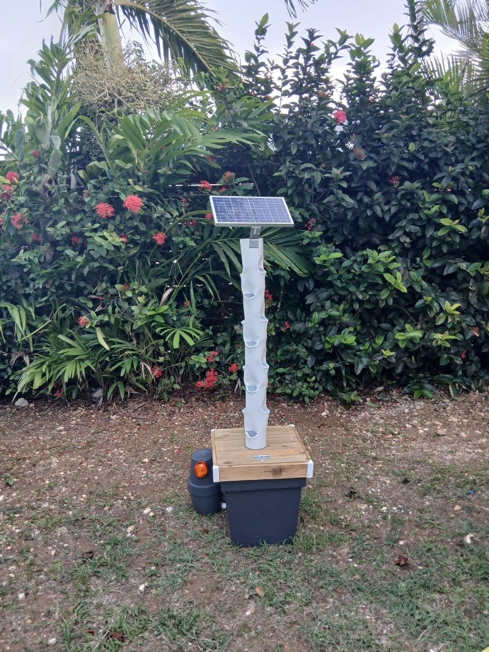 Tour de potager vertical Cultivez Facilement installée en extérieur, avec panneau solaire, devant une haie fleurie